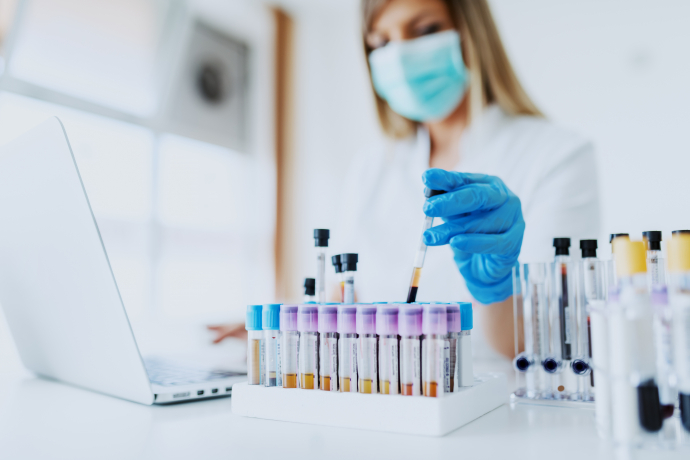 Kobieta znajdujaca się w laboratorium siedzi w maseczce przy stole, na którym znajduje się laptop i pojemnik z próbkami krwii. Kobieta pisząc na klawiaturze odkłada próbówkę z krwią do pojemnika.